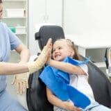 Little patient girl sitting in a chair gives a high five to a pediatric endodontics after dental treatment at a clinic with modern equipment.